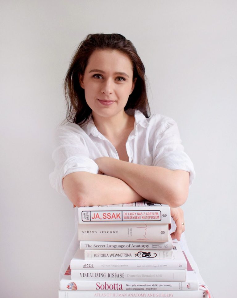 Marta Pawelec, ilustratorka medyczna, opiera ręce na stosie książek o anatomii i medycynie